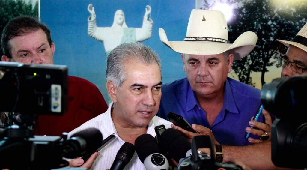 O governador Reinaldo Azambuja, durante entrevista nesta sexta-feira em Três Lagoas, observado pelo prefeito Angelo Guerreiro, de chapéu, logo atrás (Foto: Chico Ribeiro)
